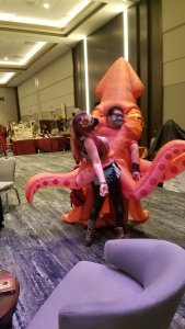 Masked person held by tentacles of person in giant orange inflatable squid costume.
