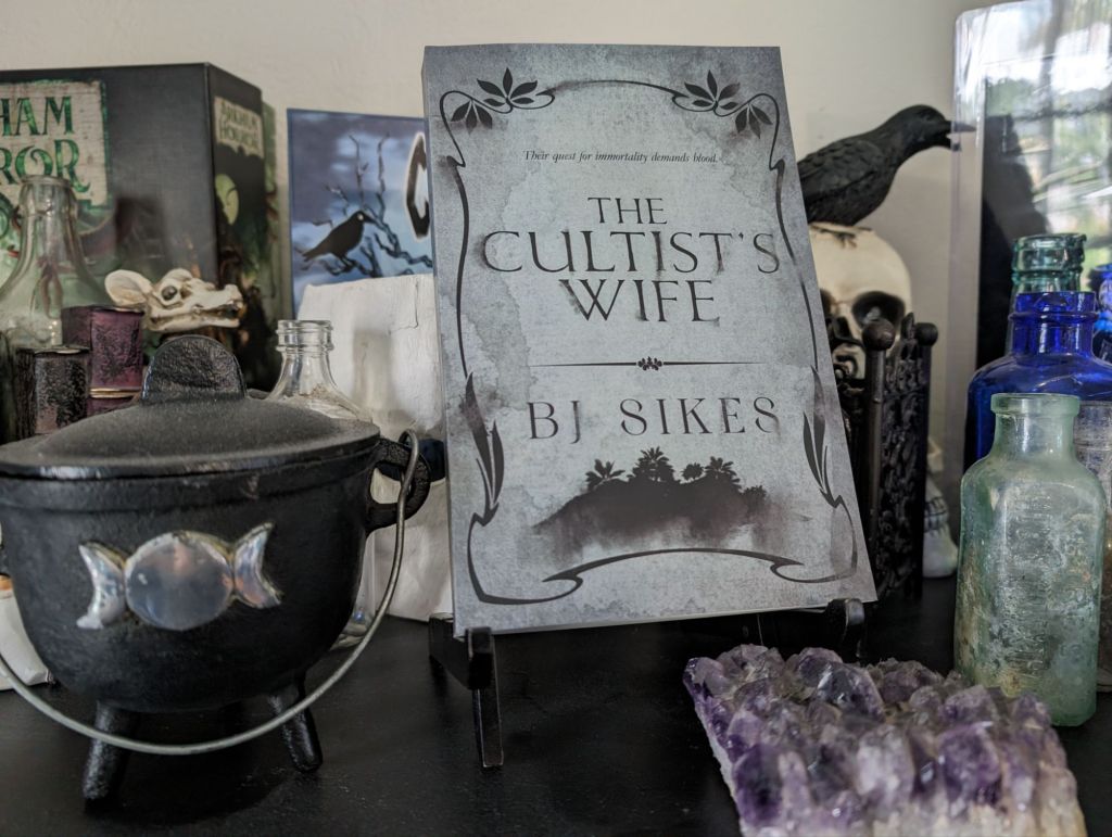 Paperbook of the Cultist's Wife on a shelf with a witch's cauldron, antique glass bottles, a crow statue, and an amethyst cluster.